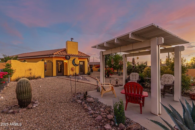 back of house at dusk with a pergola, a patio, stucco siding, a chimney, and a tiled roof