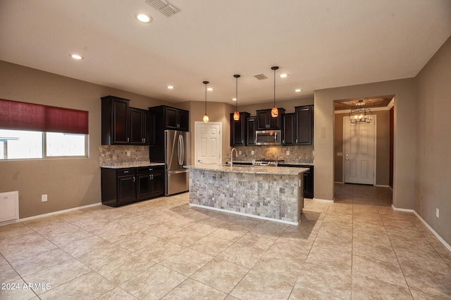 kitchen featuring a kitchen island with sink, hanging light fixtures, backsplash, stainless steel appliances, and light stone counters