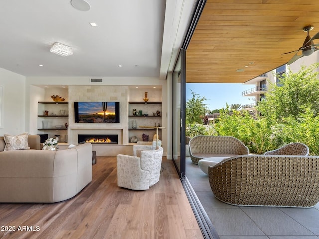 living room with wood finished floors, a ceiling fan, a lit fireplace, and built in features
