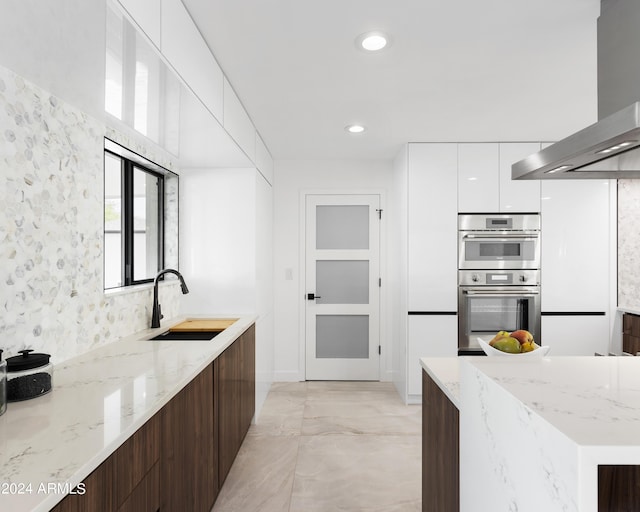 kitchen with white cabinetry, stainless steel double oven, dark brown cabinetry, sink, and wall chimney range hood