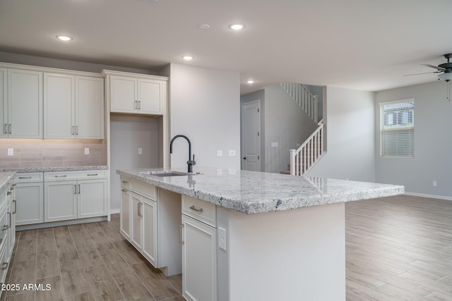 kitchen with a center island with sink, white cabinets, light wood-style flooring, light stone countertops, and a sink