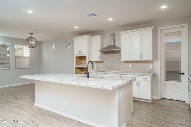 kitchen with white cabinetry, a center island with sink, wall chimney range hood, and a sink
