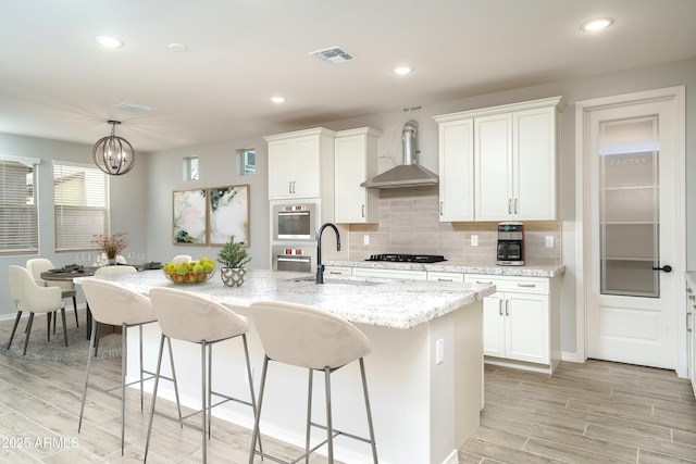 kitchen with a kitchen island with sink, a sink, visible vents, white cabinets, and wall chimney exhaust hood