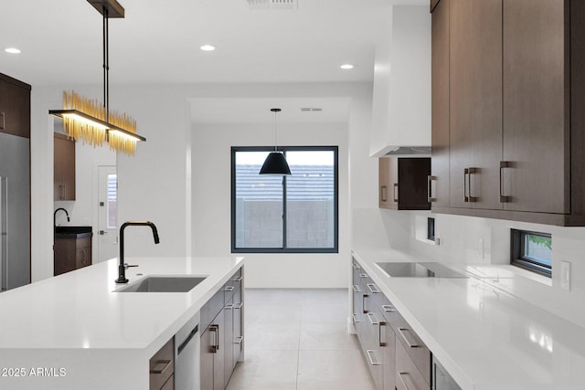 kitchen featuring wall chimney exhaust hood, sink, pendant lighting, stainless steel appliances, and a kitchen island with sink