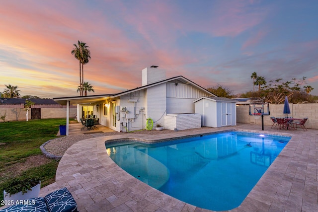 pool at dusk with a fenced backyard, an outdoor structure, outdoor dining space, a storage unit, and a patio