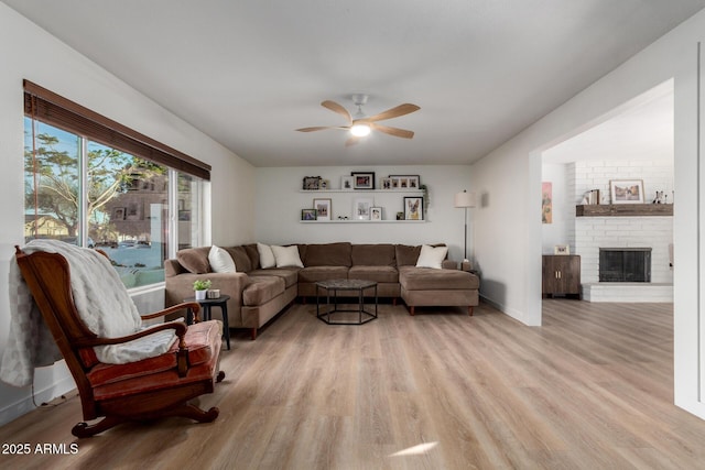 living room featuring light wood-style flooring, a brick fireplace, baseboards, and a ceiling fan