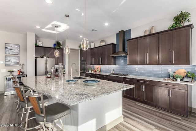 kitchen featuring tasteful backsplash, appliances with stainless steel finishes, a sink, wall chimney range hood, and a large island with sink