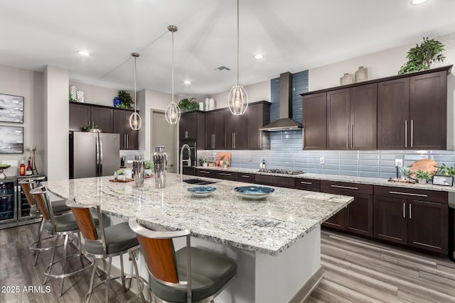kitchen featuring a large island with sink, decorative backsplash, a sink, appliances with stainless steel finishes, and wall chimney exhaust hood