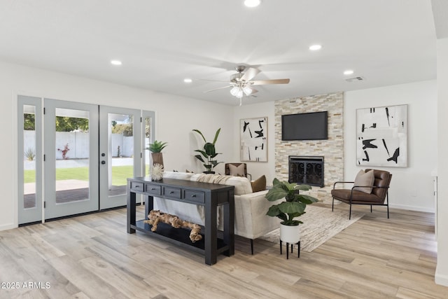 living room featuring ceiling fan, a stone fireplace, light hardwood / wood-style floors, and french doors