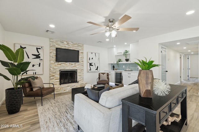 living room with wine cooler, a stone fireplace, ceiling fan, and light wood-type flooring