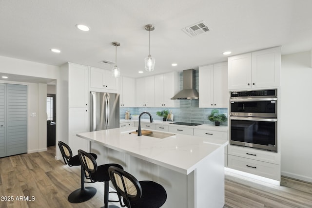 kitchen featuring appliances with stainless steel finishes, an island with sink, sink, white cabinets, and wall chimney range hood