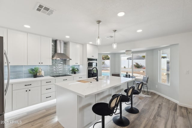 kitchen with a kitchen island with sink, wall chimney range hood, white cabinetry, and hanging light fixtures