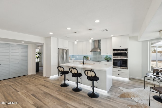 kitchen featuring white cabinetry, appliances with stainless steel finishes, a kitchen breakfast bar, a kitchen island with sink, and wall chimney range hood