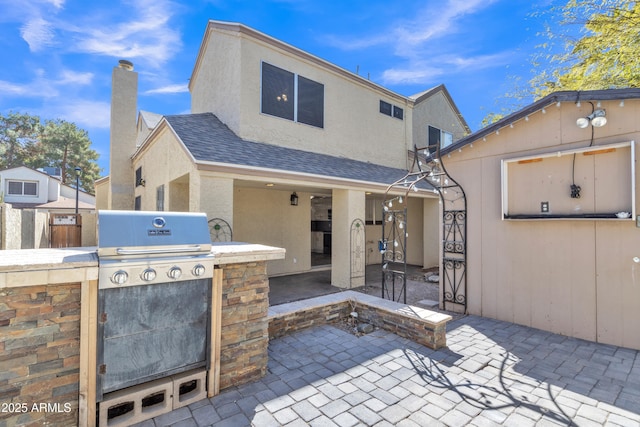 back of house with an outdoor kitchen, a shingled roof, stucco siding, a chimney, and a patio area