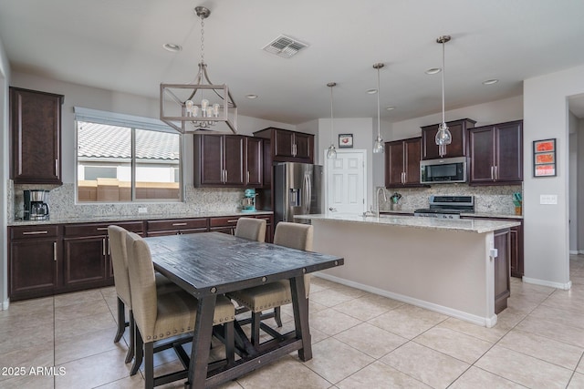 kitchen with dark brown cabinetry, a center island with sink, appliances with stainless steel finishes, pendant lighting, and light stone countertops