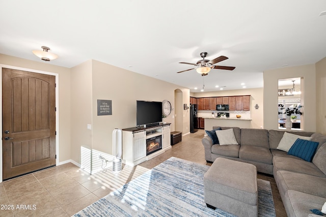 living room with light tile patterned floors, baseboards, ceiling fan with notable chandelier, and a glass covered fireplace