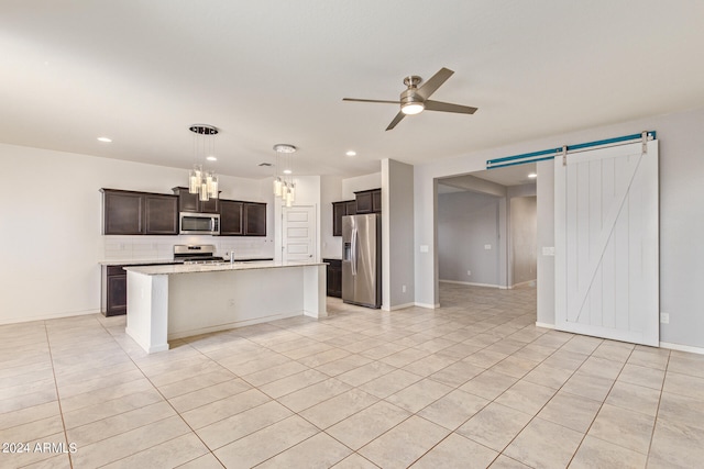 kitchen with a barn door, ceiling fan with notable chandelier, light stone counters, stainless steel appliances, and a center island with sink