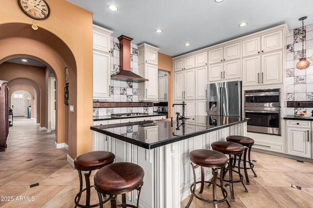 kitchen with a center island with sink, arched walkways, appliances with stainless steel finishes, a kitchen breakfast bar, and wall chimney range hood