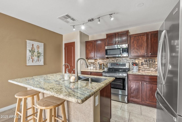 kitchen featuring appliances with stainless steel finishes, a kitchen island with sink, tasteful backsplash, light stone countertops, and a kitchen bar