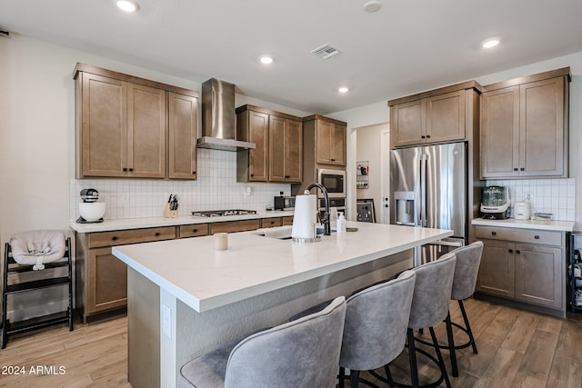 kitchen with light hardwood / wood-style floors, wall chimney range hood, an island with sink, and stainless steel appliances