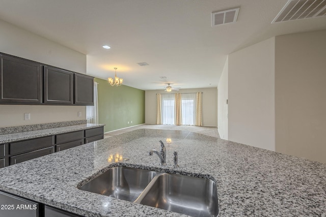 kitchen with sink, hanging light fixtures, dark brown cabinets, light stone counters, and ceiling fan with notable chandelier