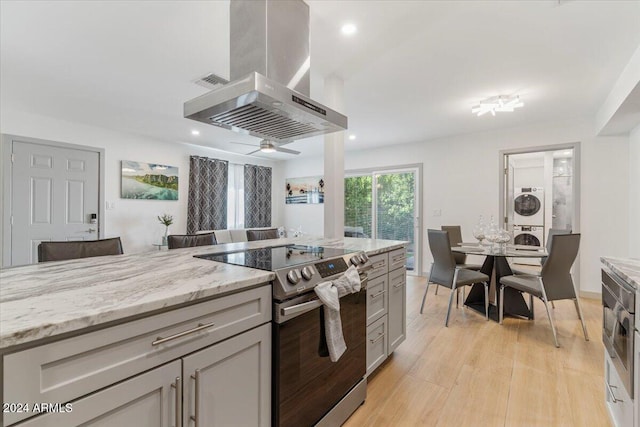 kitchen featuring stainless steel electric range, stacked washer and clothes dryer, ceiling fan, light stone counters, and island exhaust hood