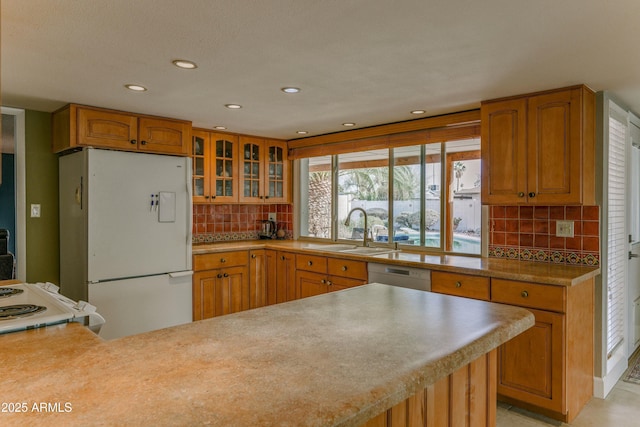 kitchen featuring tasteful backsplash, glass insert cabinets, brown cabinetry, a sink, and white appliances