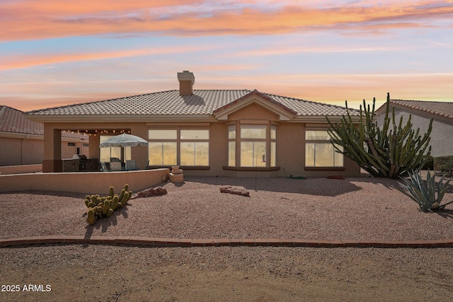 back of house at dusk featuring a patio area, a tile roof, a chimney, and stucco siding