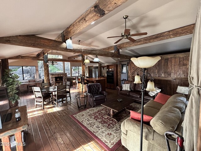 living room with ceiling fan, lofted ceiling with beams, dark hardwood / wood-style floors, a stone fireplace, and wood walls