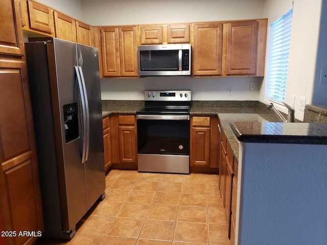 kitchen with light tile patterned floors, brown cabinetry, a peninsula, appliances with stainless steel finishes, and dark countertops