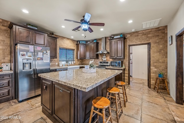 kitchen with appliances with stainless steel finishes, a kitchen bar, a center island, dark brown cabinetry, and wall chimney exhaust hood