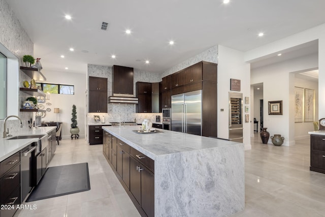 kitchen featuring wall chimney range hood, built in appliances, backsplash, a large island, and sink