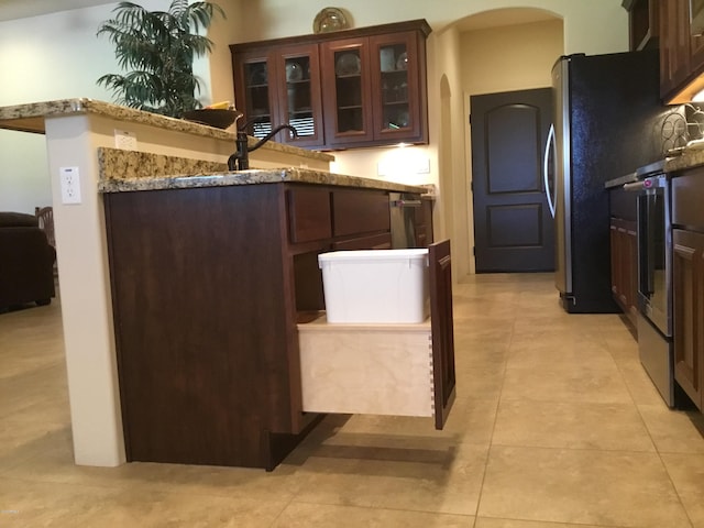 kitchen with light tile patterned floors, arched walkways, glass insert cabinets, light stone counters, and freestanding refrigerator