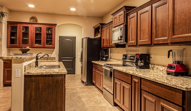 kitchen featuring arched walkways, a sink, appliances with stainless steel finishes, light stone countertops, and glass insert cabinets