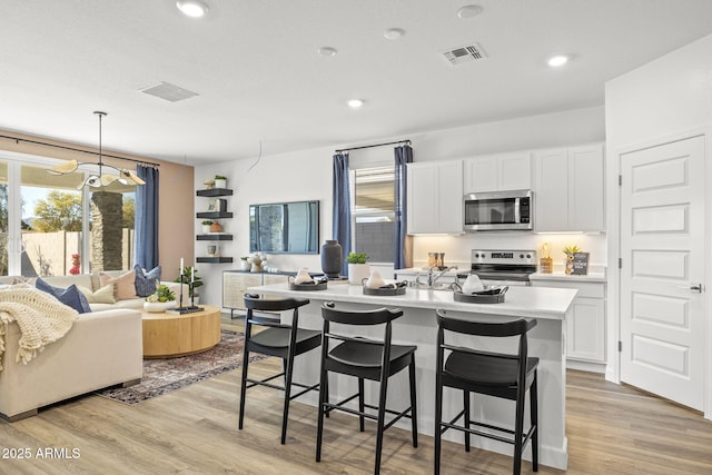 kitchen with light hardwood / wood-style flooring, hanging light fixtures, stainless steel appliances, white cabinets, and a center island with sink