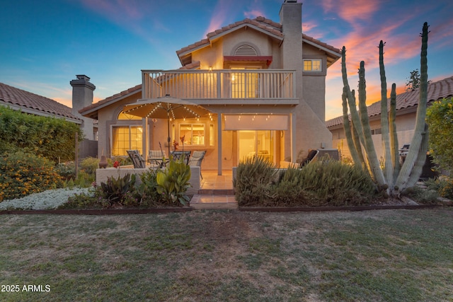 back of house at dusk featuring a tiled roof, a chimney, stucco siding, and a balcony