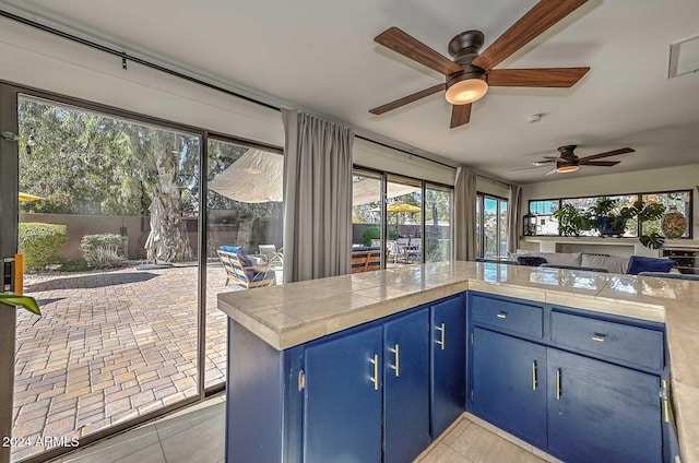 kitchen with kitchen peninsula, ceiling fan, blue cabinetry, light tile patterned floors, and tile counters