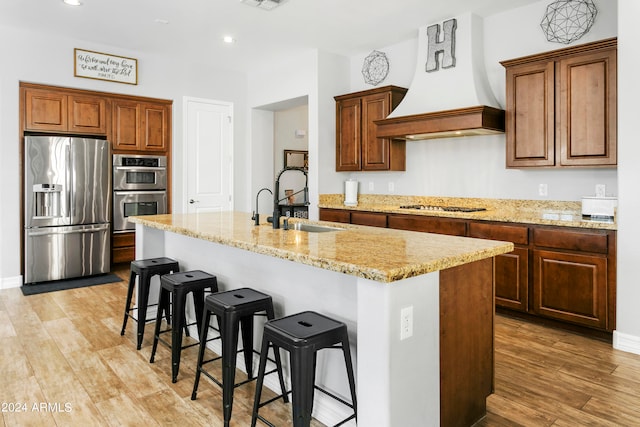 kitchen featuring light wood-type flooring, appliances with stainless steel finishes, a center island with sink, and custom range hood