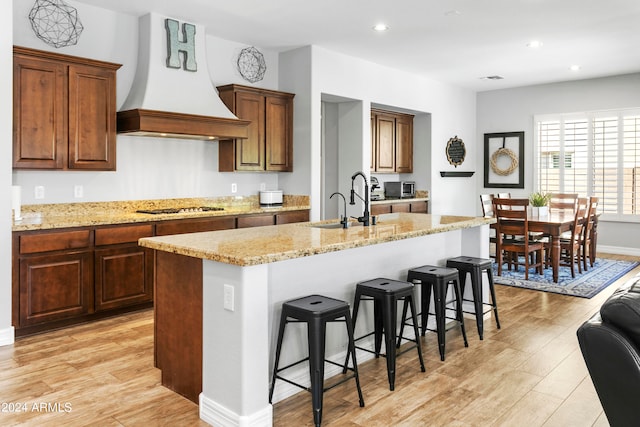 kitchen with custom exhaust hood, stainless steel gas cooktop, a kitchen bar, a center island with sink, and light hardwood / wood-style floors