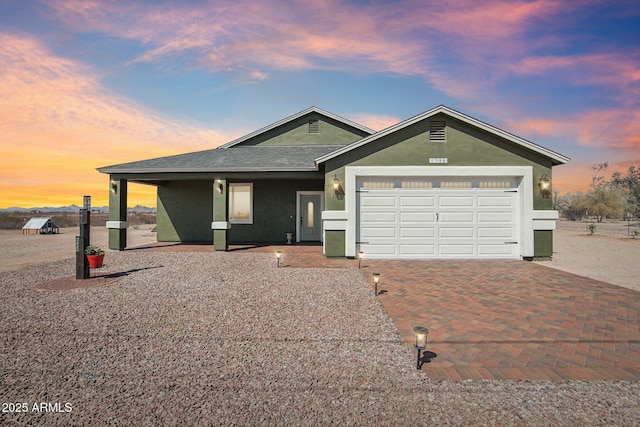 ranch-style house with a shingled roof, decorative driveway, an attached garage, and stucco siding