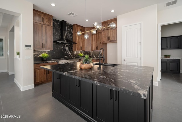 kitchen with wall chimney exhaust hood, a sink, backsplash, a kitchen island with sink, and brown cabinets