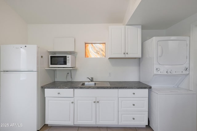 kitchen with dark countertops, white cabinets, a sink, stacked washing maching and dryer, and white appliances