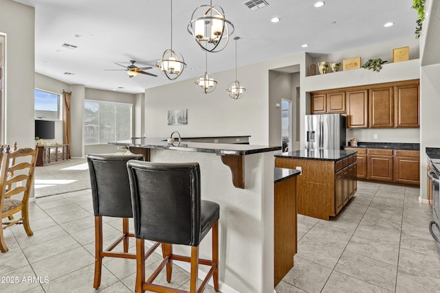 kitchen featuring brown cabinetry, dark countertops, a kitchen breakfast bar, a kitchen island with sink, and stainless steel refrigerator with ice dispenser