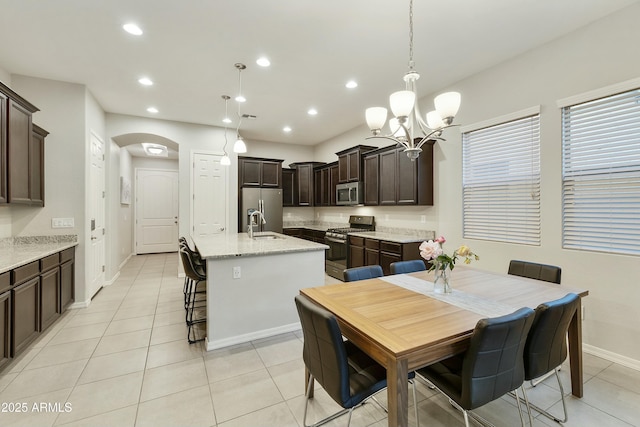 kitchen with light stone countertops, dark brown cabinets, stainless steel appliances, a kitchen island with sink, and hanging light fixtures