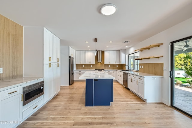 kitchen featuring white cabinets, appliances with stainless steel finishes, a kitchen island, decorative light fixtures, and wall chimney range hood