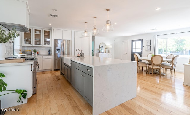 kitchen featuring white cabinetry, decorative light fixtures, light wood-type flooring, appliances with stainless steel finishes, and a kitchen island with sink