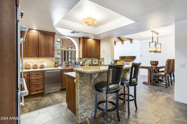 kitchen featuring decorative backsplash, a raised ceiling, a breakfast bar area, stone tile flooring, and stainless steel dishwasher