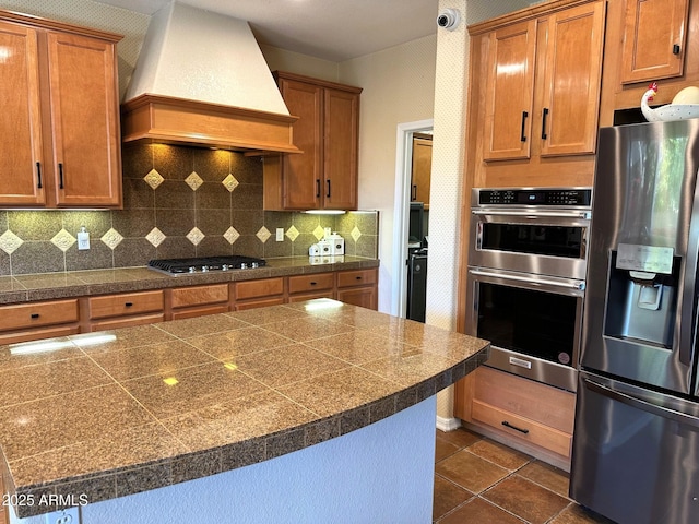 kitchen featuring premium range hood, dark tile patterned flooring, backsplash, appliances with stainless steel finishes, and tile counters