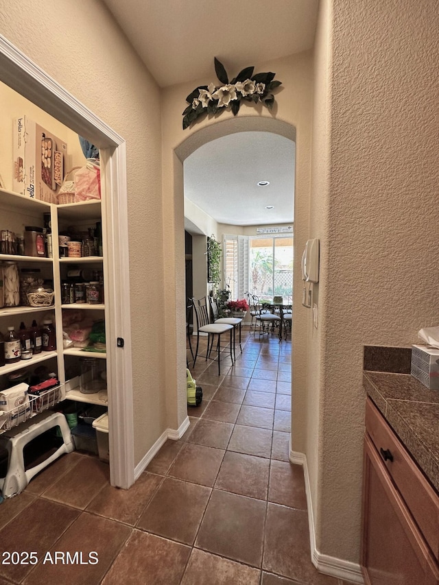 hall featuring dark tile patterned floors, a textured wall, arched walkways, and baseboards