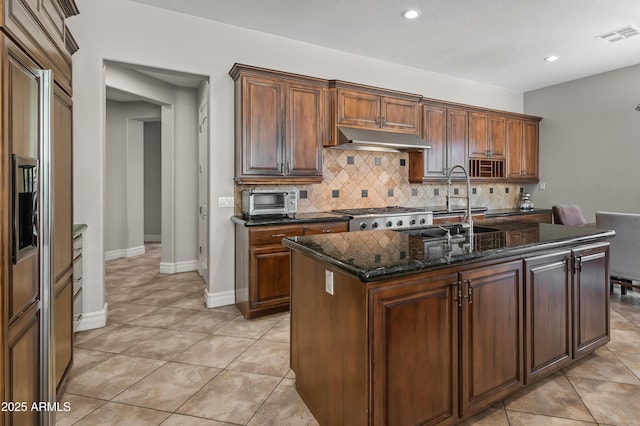 kitchen featuring visible vents, under cabinet range hood, dark stone countertops, a sink, and decorative backsplash